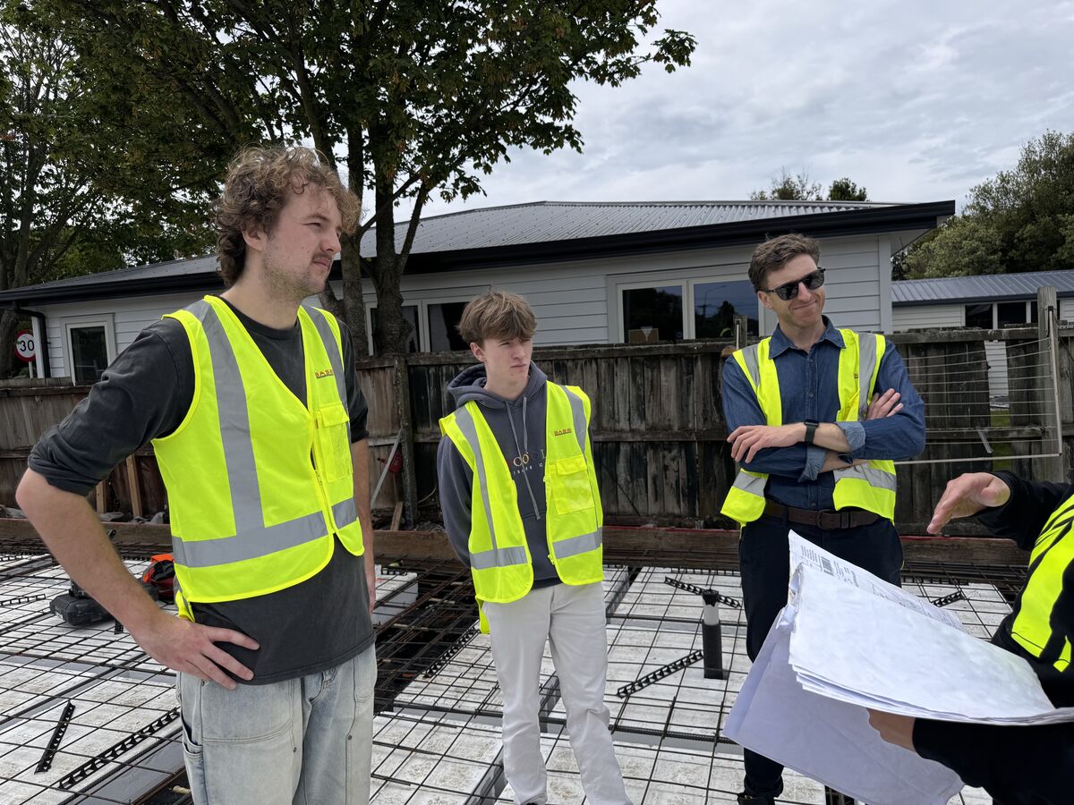 Three men wearing yellow safety vests at a construction site reviewing blueprints.
