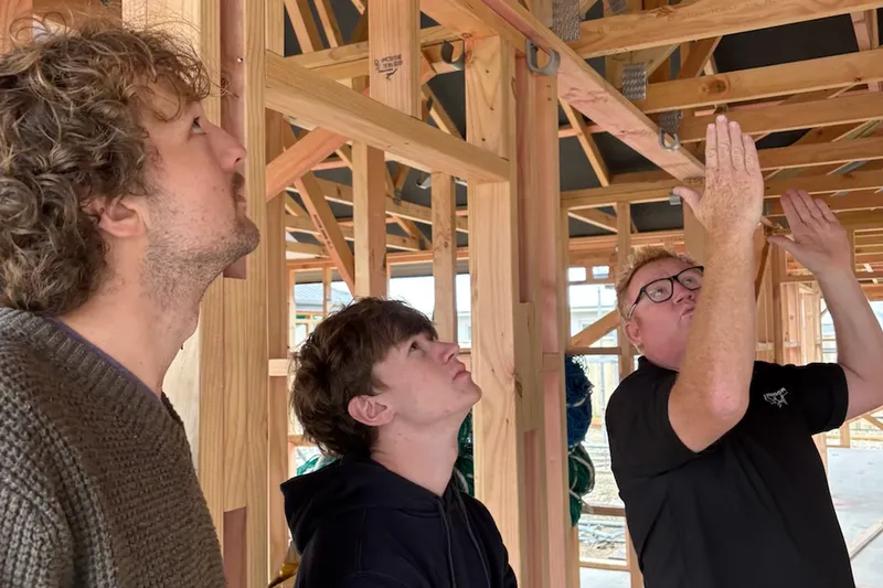 Three men inside a wooden house frame, one explaining something while pointing upwards.